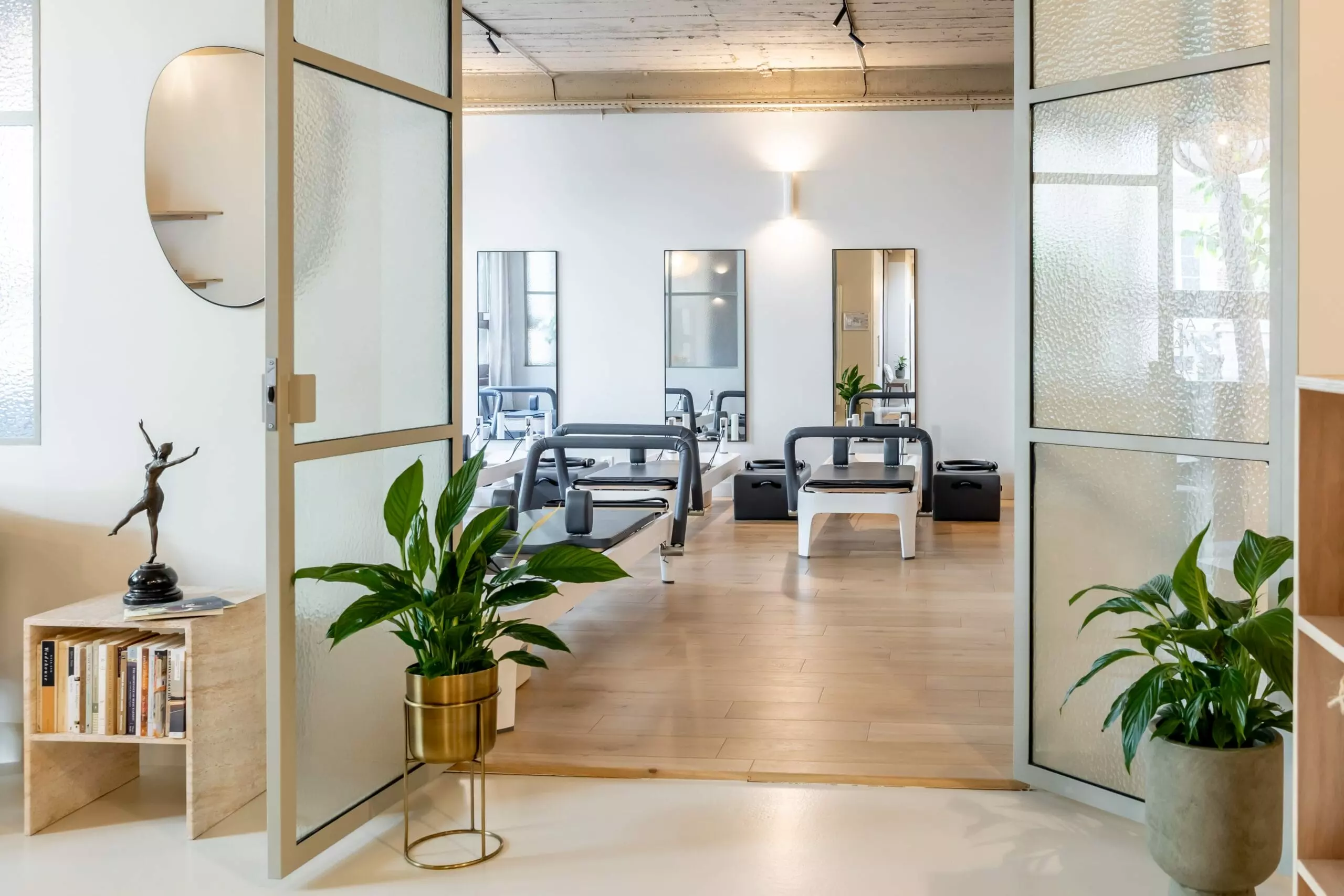 Salle de Pilates moderne depuis l'exterieur équipée de reformers alignés, avec un sol en bois clair, des murs blancs, une décoration végétale et un éclairage chaleureux.