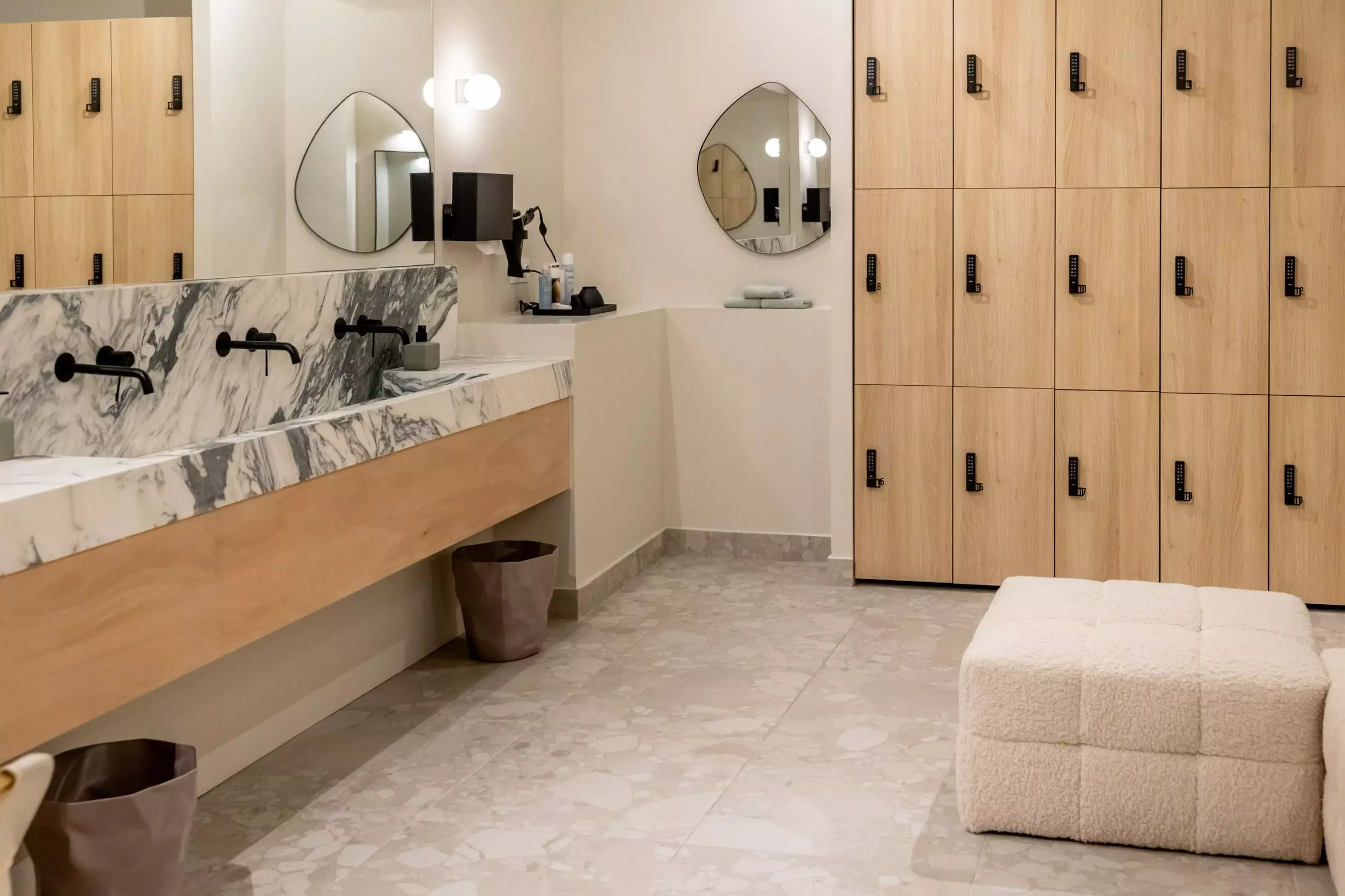 Espace vestiaire élégant avec casiers en bois clair, banquette moelleuse, rideaux beiges, miroir mural, décoration artistique minimaliste et lavabos en marbre mis en valeur.