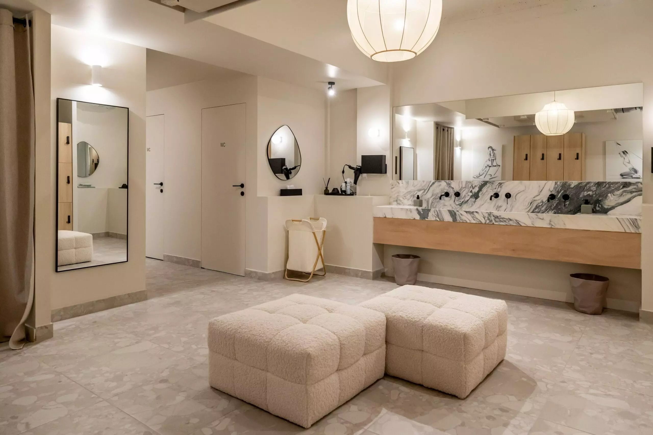 Espace vestiaire élégant avec casiers en bois clair, banquette moelleuse, rideaux beiges, miroir mural et décoration artistique minimaliste.