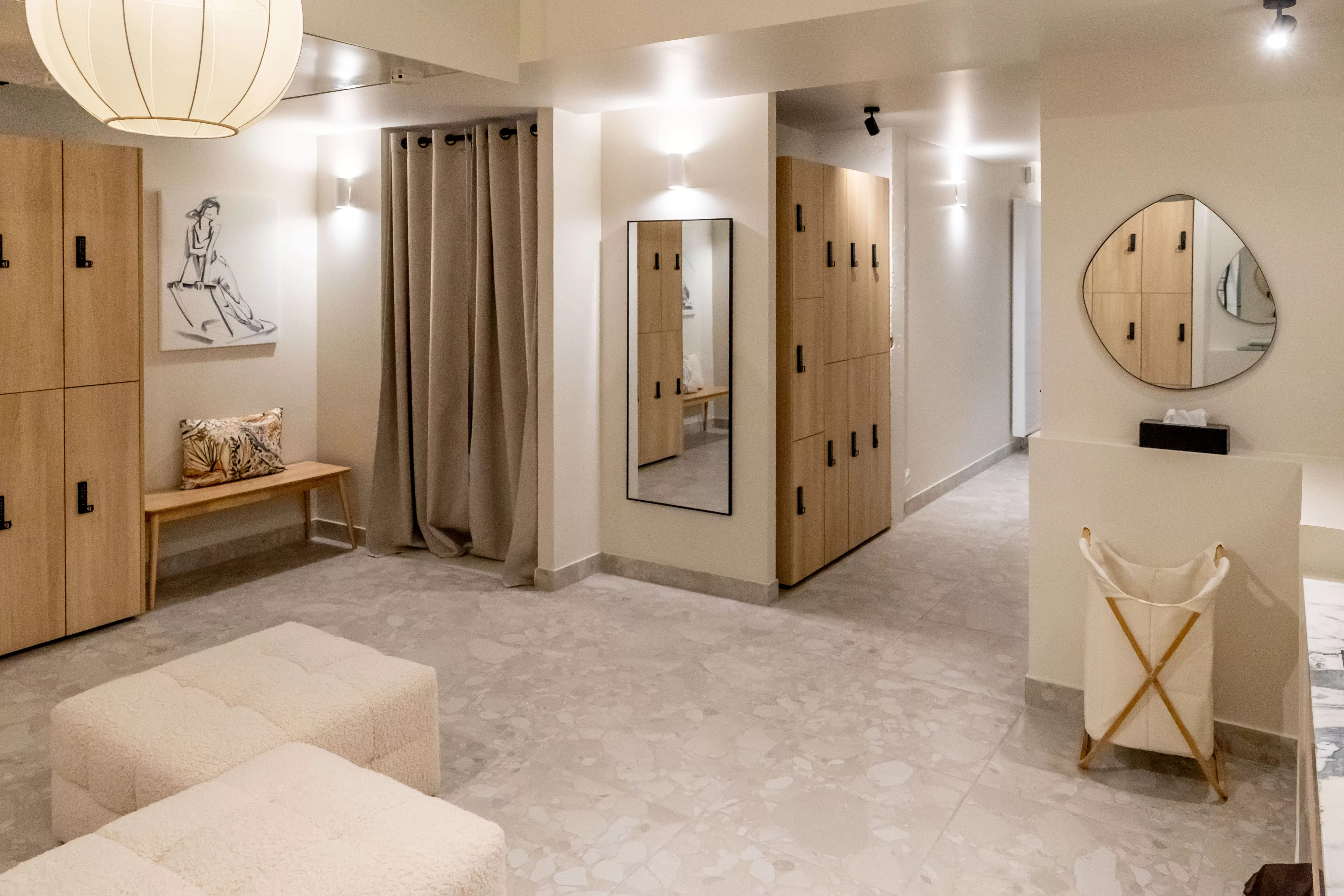 Espace vestiaire élégant avec casiers en bois clair, banquette moelleuse, rideaux beiges, miroir mural et décoration artistique minimaliste.