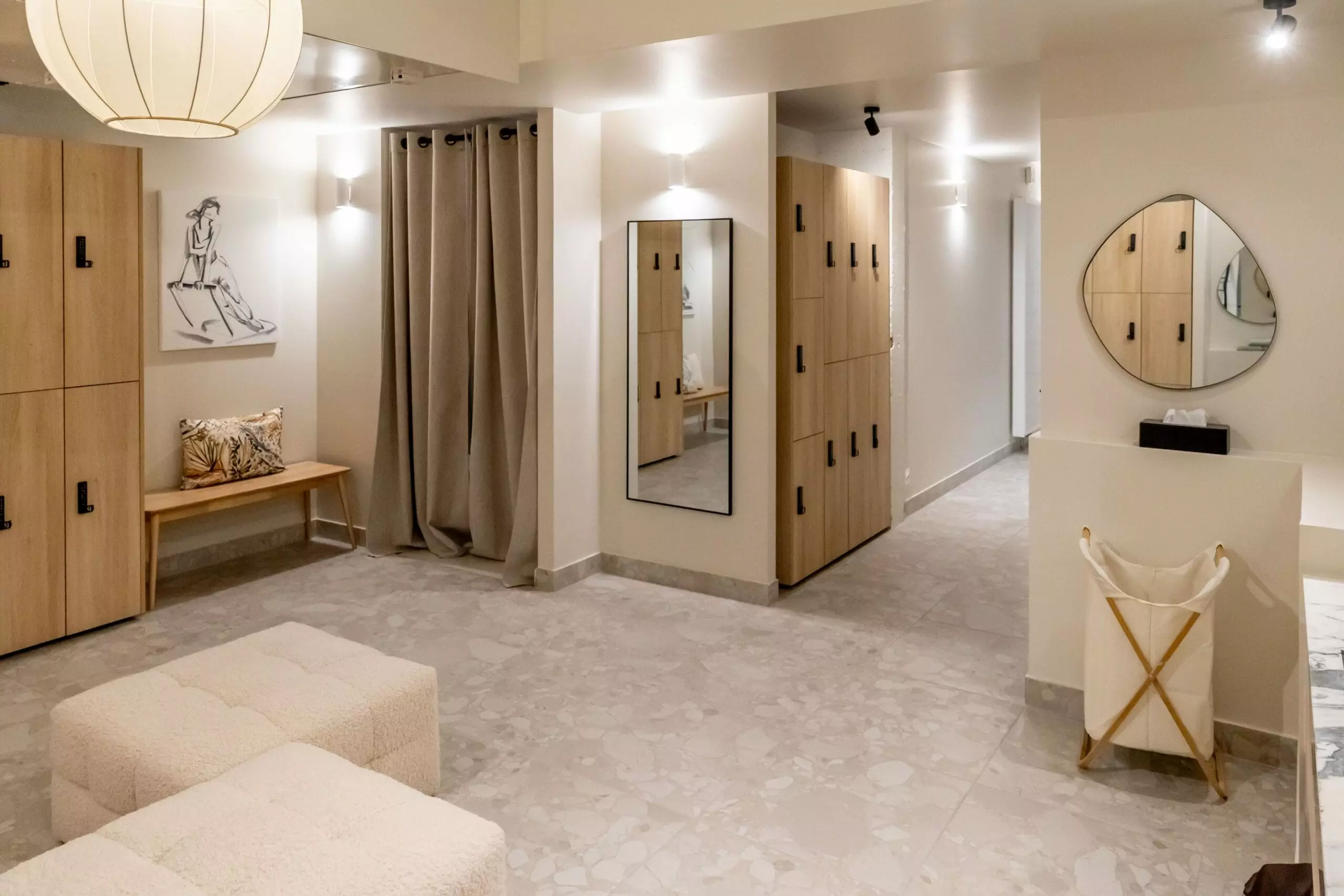Espace vestiaire élégant avec casiers en bois clair, banquette moelleuse, rideaux beiges, miroir mural et décoration artistique minimaliste.