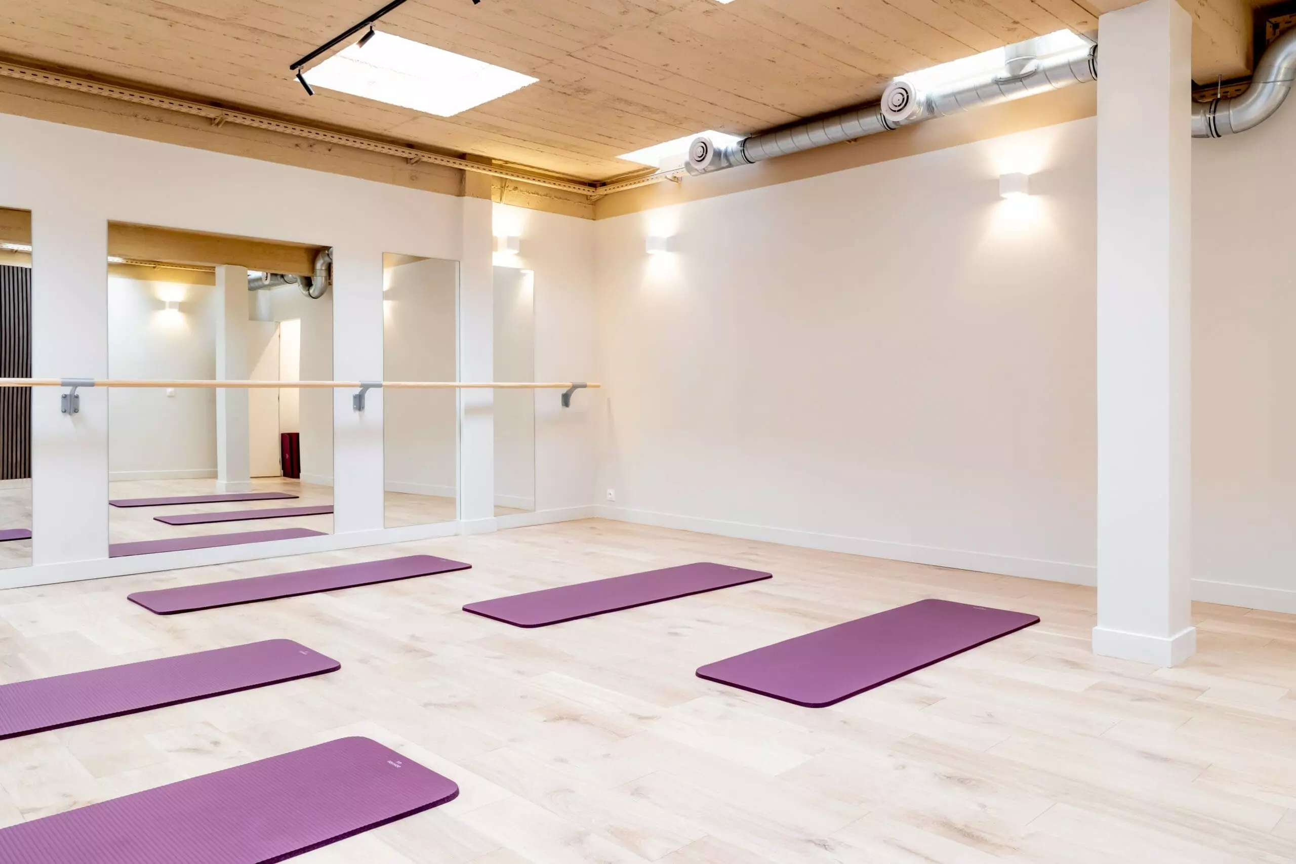 Salle de sport lumineuse avec parquet clair, murs blancs, miroirs, barres de danse et tapis de yoga violets alignés au sol.