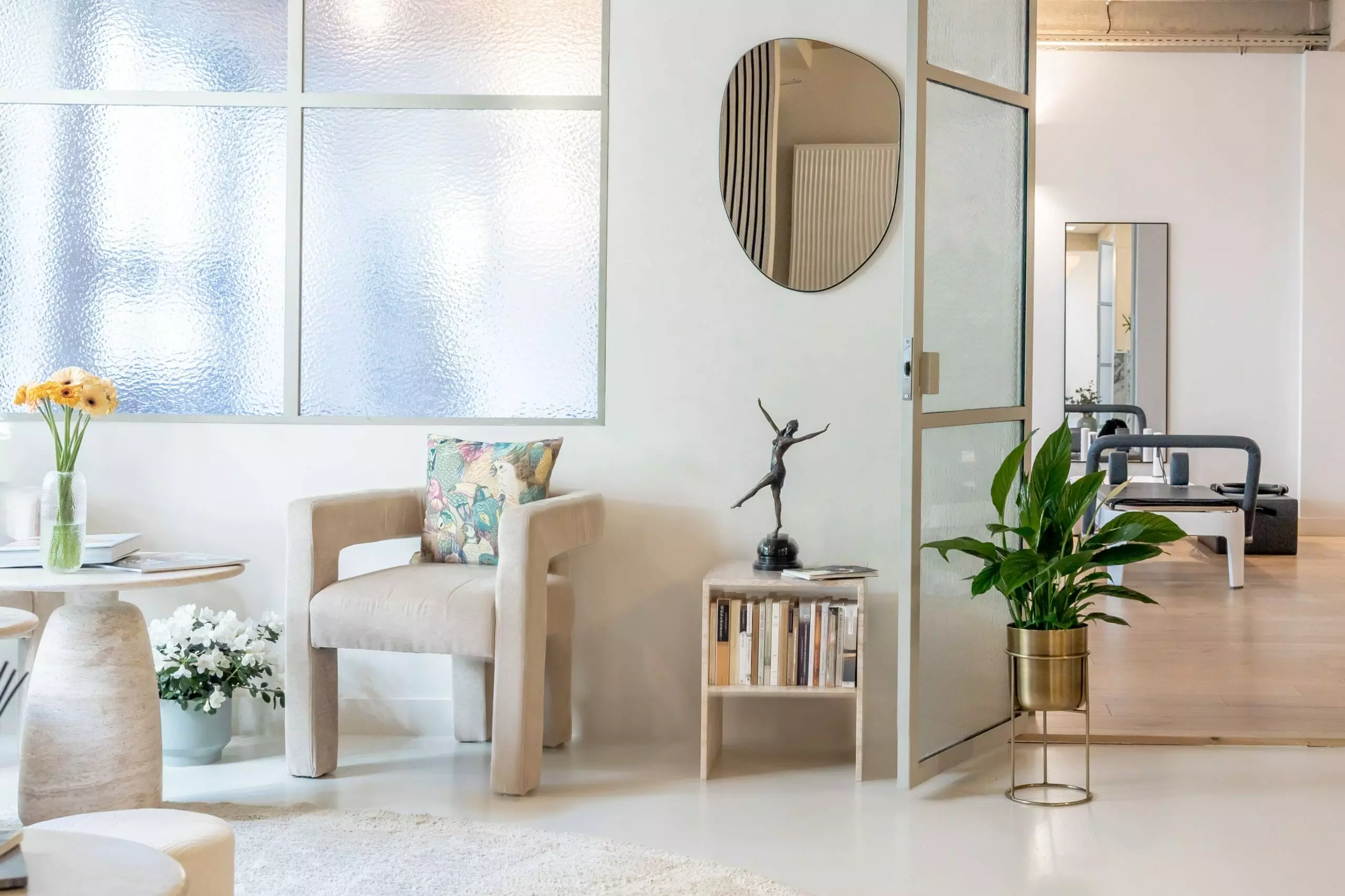 Une pièce moderne et lumineuse avec un fauteuil beige, une petite table, des plantes en pot, un miroir rond et une paroi en verre dépoli. En arrière-plan, du matériel de sport est visible