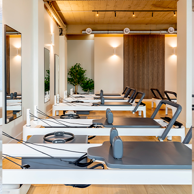 Salle de Pilates moderne équipée de reformers alignés, avec un sol en bois clair, des murs blancs, une décoration végétale et un éclairage chaleureux.