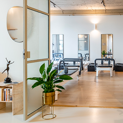 "Entrée moderne d'une salle de Pilates Reformer, avec un ambiance minimaliste et lumineuse. Un petit bureau porte des livres et une statue de danseuse, accompagné d'une plante en pot. À gauche, un fauteuil beige invite à la détente, et une porte vitrée mène à une autre pièce avec du matériel de sport.