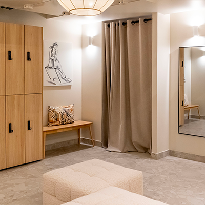 Espace vestiaire élégant avec casiers en bois clair, banquette moelleuse, rideaux beiges, miroir mural et décoration artistique minimaliste.