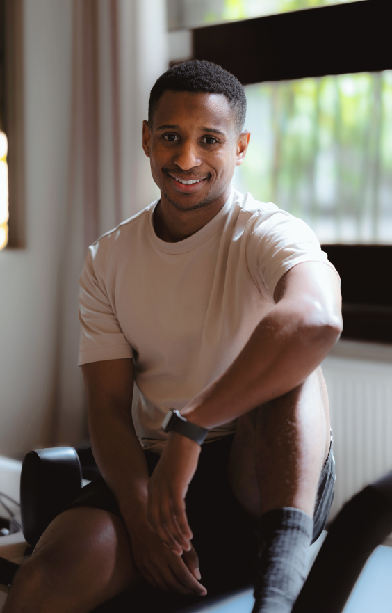Un homme vêtu d'un t-shirt beige clair et d'un short foncé est assis sur une chaise à l'intérieur, souriant à la caméra. Il a les cheveux courts, porte une montre connectée et la lumière du soleil entre par une fenêtre avec des stores derrière lui et un Pilates Reformer à proximité.
