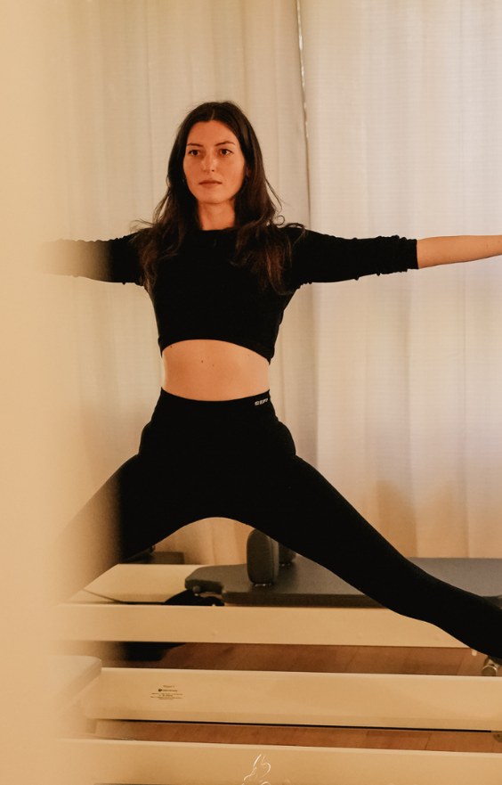 Une femme en tenue de sport noire effectue une pose d'étirement sur une machine Pilates Reformer à l'intérieur, les bras étendus et l'expression concentrée.