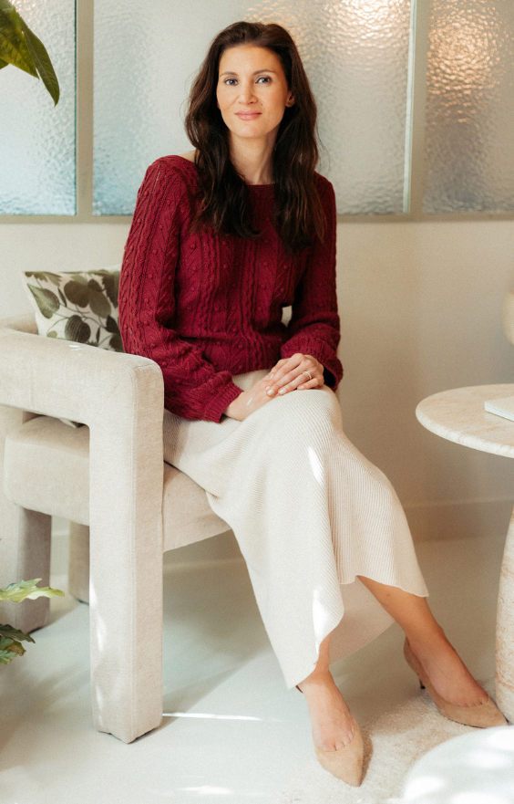 Une femme assise pose calmement dans un intérieur lumineux et élégant. Elle a la peau claire, des yeux foncés et de longs cheveux bruns tombant librement sur ses épaules. Elle porte un pull en maille rouge bordeaux et une jupe longue beige en tissu fluide, ainsi que des chaussures beiges à talons bas. Elle est installée dans un fauteuil beige clair, à côté d'une table ronde assortie, avec un coussin à motif de feuilles derrière elle. L’arrière-plan est composé de panneaux de verre dépoli, laissant passer une lumière douce et diffuse. Son expression est sereine et accueillante.