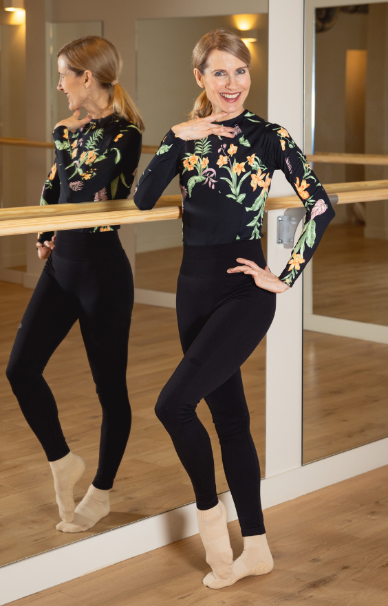Une femme souriante se tient dans un studio de danse spacieux et lumineux, avec un sol en bois clair et de grands miroirs. Elle pose élégamment près d'une barre de danse, une main posée sur la taille et l'autre sous son menton. Elle a la peau claire, les yeux bleus et les cheveux blonds attachés en une queue de cheval soignée. Elle porte une tenue noire moulante : un haut à manches longues orné de motifs floraux colorés, un legging noir et des chaussettes beiges. Son reflet est clairement visible dans le miroir derrière elle, ajoutant de la profondeur et de la lumière à la scène.