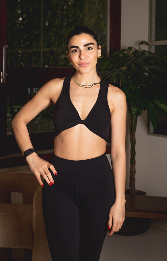 Une femme vêtue d'une brassière de sport noire et d'un legging taille haute se tient à l'intérieur, souriant à la caméra après une séance de Pilates. Ses cheveux noirs sont tirés en arrière et elle porte un collier, une montre et du vernis à ongles rouge. Une plante et une chaise sont visibles en arrière-plan.