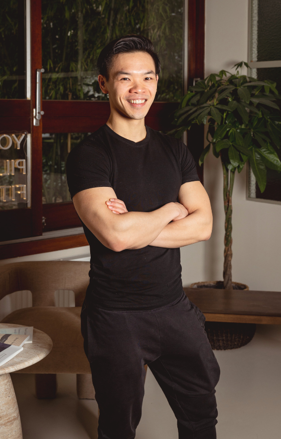 Un homme souriant, vêtu d'un t-shirt et d'un pantalon noirs, se tient à l'intérieur, les bras croisés, comme un professeur de Pilates. Derrière lui se trouvent une plante en pot, un banc en bois et une table en marbre avec des papiers. La lumière naturelle entre par la fenêtre.