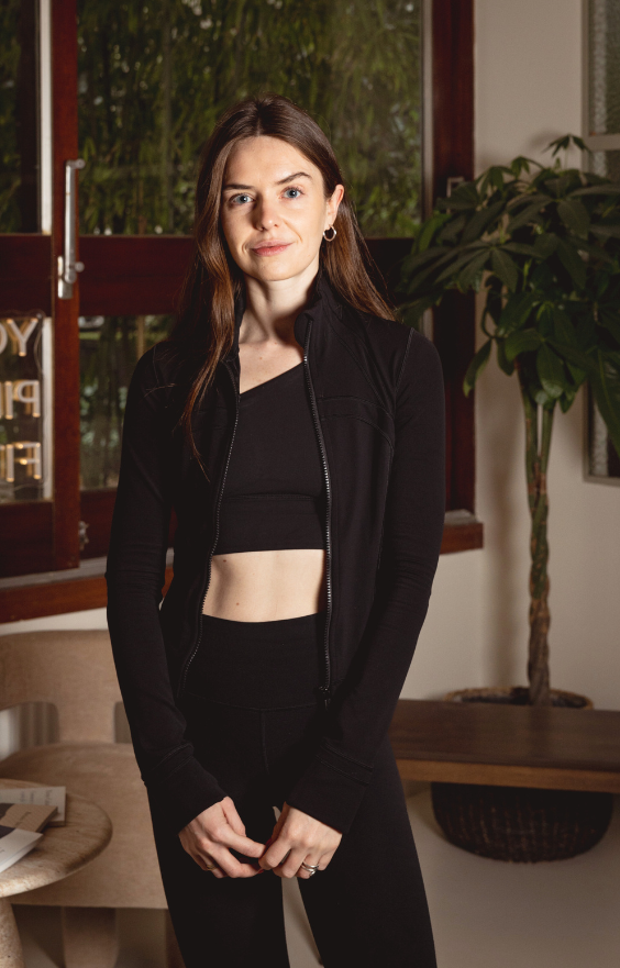 Femme en position fière, les mains sur les hanches, professeur de Yoga vêtue de noir, cheveux bruns et lèvres légèrement rouges. En arrière-plan, l'entrée du studio de Pilates et Yoga Aja House à Paris avec une plante.