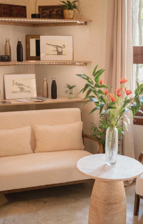 Coin café chaleureux dans un studio de Pilates, décoré avec des œuvres d'art inspirées du Pilates sur les étagères murales. L’espace est aménagé avec un canapé confortable, une table ronde en bois et un vase de fleurs fraîches, créant une ambiance apaisante et accueillante.