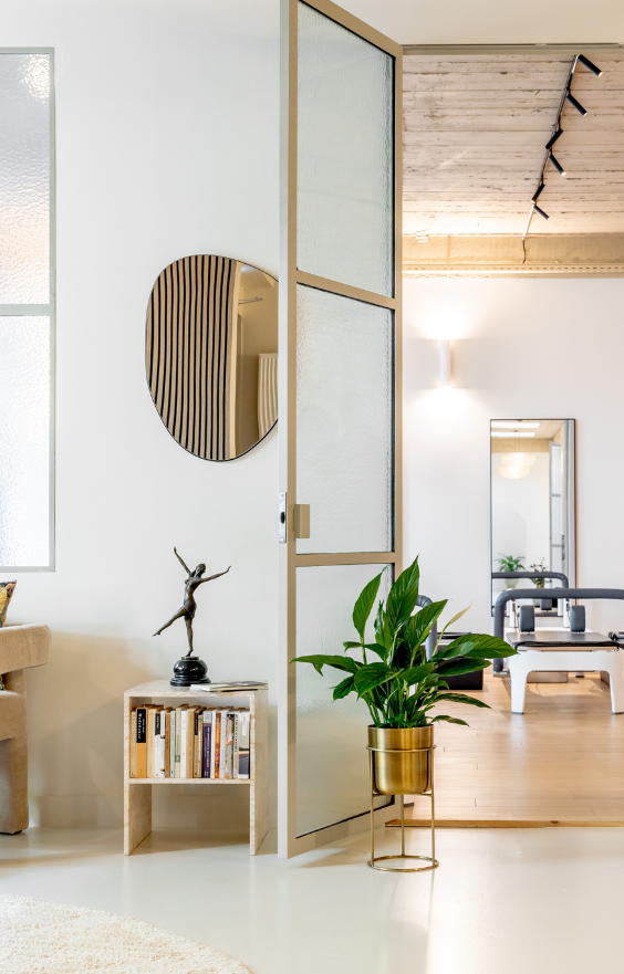 Une pièce moderne et lumineuse avec un fauteuil beige, une petite table, des plantes en pot, un miroir rond et une paroi en verre dépoli. En arrière-plan, du matériel de sport est visible
