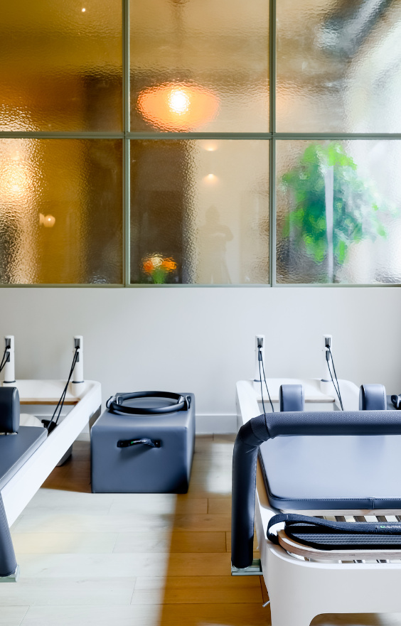 Salle de Pilates moderne équipée de reformers alignés, avec un sol en bois clair, des murs blancs avec une verrière, une décoration végétale et un éclairage naturel.