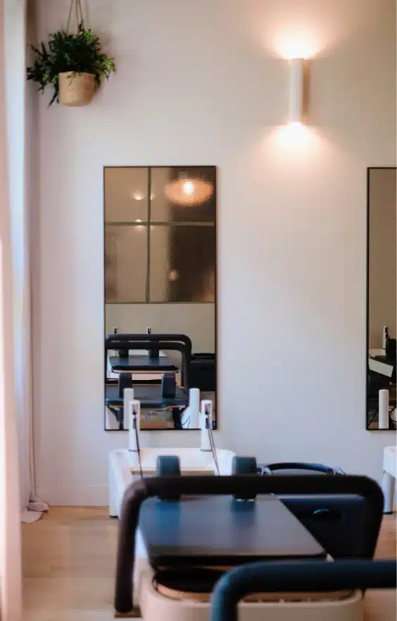 Une pièce moderne et lumineuse avec une plante verte suspendue, un miroir rond et une machine de Pilates Reformer