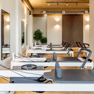 Salle de Pilates moderne équipée de reformers alignés, avec un sol en bois clair, des murs blancs, une décoration végétale et un éclairage chaleureux.