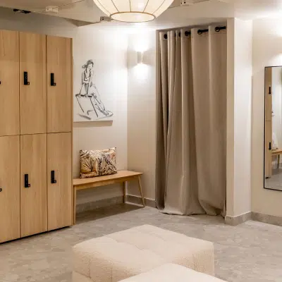 Espace vestiaire élégant avec casiers en bois clair, banquette moelleuse, rideaux beiges, miroir mural et décoration artistique minimaliste.